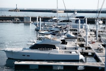 A marina with several yachts docked side by side, showcasing a tranquil harbor setting. The water is calm, and the boats are moored at wooden docks. A breakwater and lighthouse are visible in the background, with a clear sky overhead.