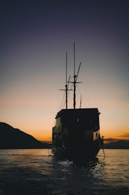 A large sailboat is silhouetted against a vibrant sunset. The sky is painted with gradients of orange and purple, creating a serene and picturesque scene. The water reflects the colors of the sky, enhancing the tranquil atmosphere. A mountainous landscape is visible in the background.