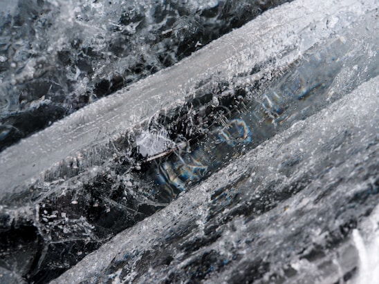 Crystal-like structures of ice with intricate patterns and textures. The ice appears clear with small air bubbles and fissures throughout, creating a complex, layered visual effect.
