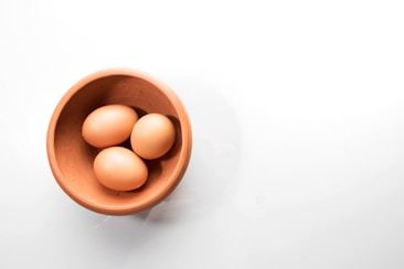 A wooden bowl contains three brown eggs against a minimalist white background.