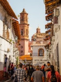 A bustling street scene in a historic town with a tall, ornate cathedral tower prominently in the background. The street is lined with white buildings featuring wooden balconies and tile roofs. A diverse group of people, including individuals walking and engaging in various activities, fills the street. A sign for 'Hotel Melendez' and a 'Restaurant' is visible on one of the buildings.