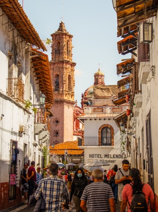A bustling street scene in a historic town with a tall, ornate cathedral tower prominently in the background. The street is lined with white buildings featuring wooden balconies and tile roofs. A diverse group of people, including individuals walking and engaging in various activities, fills the street. A sign for 'Hotel Melendez' and a 'Restaurant' is visible on one of the buildings.