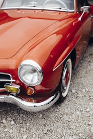 A classic car with a shiny red exterior and chrome detailing is parked on a gravel surface. The car features round headlights and a sleek, curving body design, typical of vintage automobiles.