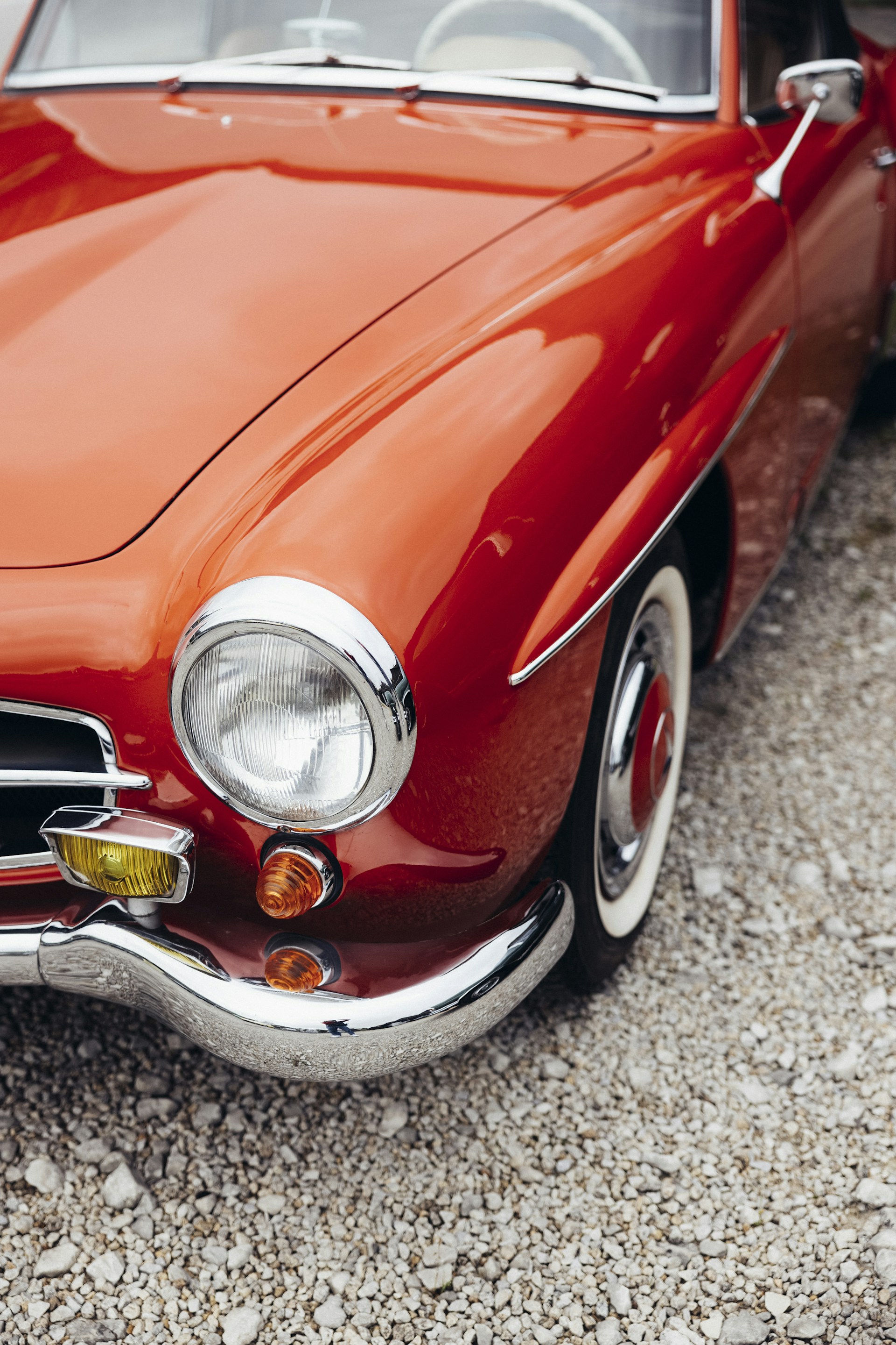 A classic car with a shiny red exterior and chrome detailing is parked on a gravel surface. The car features round headlights and a sleek, curving body design, typical of vintage automobiles.