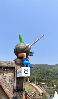 A whimsical statue of a bird-like creature with a large beak, feathered head, and colorful body stands on a rustic wooden post. The post is part of a structure that resembles a fantasy-themed entrance, adorned with stone textures and string lights. In the background, a lush green hillside and parts of a bridge are visible under a clear blue sky.