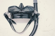 A black diving mask and snorkel set is laid out on a light surface. The mask has a wide lens area with transparent plastic and is attached to the snorkel, which is coiled around it. The surface exhibits some minor scratches and marks.