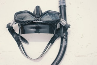 A black diving mask and snorkel set is laid out on a light surface. The mask has a wide lens area with transparent plastic and is attached to the snorkel, which is coiled around it. The surface exhibits some minor scratches and marks.