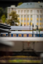 A detailed miniature scene with a model train situated in front of a large building, possibly a station or an official structure. The train is blue with lighted windows, and the scene includes realistic features such as lamp posts and trees, suggesting a setting in a small town or city environment.