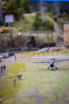 A miniature landscape scene with a small-scale airplane on a grassy field. Several model figures are present, appearing to walk and engage in activities. In the background, a forested area and utility structures are visible.