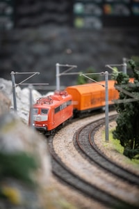 A model train with an orange locomotive travels along a curved track, surrounded by miniature greenery and overhead power lines.