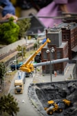 A detailed miniature scene of a construction site features a yellow crane lifting a pipe near a brick industrial building. Vehicles such as a truck and a bulldozer are positioned around a large excavation site. Model trees and street lamps line the miniature roadway. Figures of construction workers can be seen on the site, with additional background detail including a train track.
