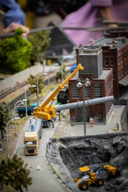 A detailed miniature scene of a construction site features a yellow crane lifting a pipe near a brick industrial building. Vehicles such as a truck and a bulldozer are positioned around a large excavation site. Model trees and street lamps line the miniature roadway. Figures of construction workers can be seen on the site, with additional background detail including a train track.