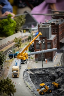 A detailed miniature scene of a construction site features a yellow crane lifting a pipe near a brick industrial building. Vehicles such as a truck and a bulldozer are positioned around a large excavation site. Model trees and street lamps line the miniature roadway. Figures of construction workers can be seen on the site, with additional background detail including a train track.