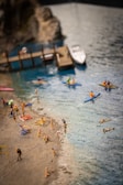 A miniature beach scene featuring small figures enjoying various activities along the sandy shore and in the water. Several figures are lounging on the beach under umbrellas, while others swim or paddle on kayaks in the blue water. A pier extends from the rocky coastline, with a small boat docked nearby.