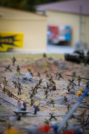 A detailed diorama depicting a wartime scene with miniature soldiers engaging in combat on a rough terrain. The background includes a blurred view of buildings and a sign, while miniature vehicles and equipment are scattered across the landscape.