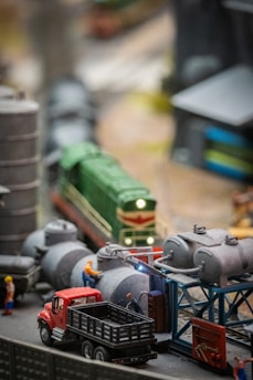 A model train set features a green locomotive on tracks, surrounded by industrial structures and equipment, including cylindrical tanks and scaffolding. Miniature figures in work attire engage with the scene, and a red truck is parked nearby.