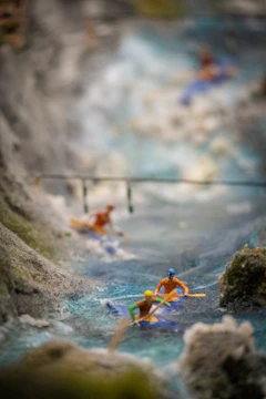 A miniature scene depicting several tiny figurines in colorful kayaks navigating a simulated river with rocky banks. The craftsmanship detail highlights the fast-flowing water effect.