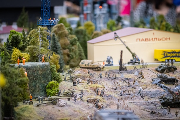 A detailed diorama depicting a wartime scene with miniature soldiers, tanks, and various military equipment spread across a terrain featuring trees and a structure labeled with Cyrillic text. Several figures in orange uniforms work on a structure in the foreground.