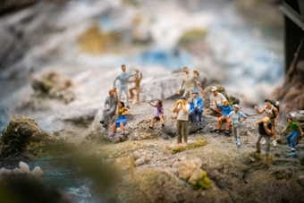 A group of miniature figurines arranged on a rocky landscape, depicting people in various poses such as sitting, standing, and interacting. The scene creates a vibrant and dynamic atmosphere, with some figures appearing to be taking photographs and others engaged in conversation.