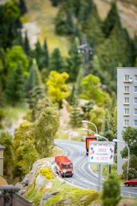 A miniature scene depicts a winding road through a lush, hilly landscape with dense green foliage. A small model of a red bus travels on the road, passing under streetlights. Part of a building and a sign are also visible alongside the road, suggesting an urban or suburban environment.