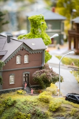 A detailed miniature model scene featuring a brick house with two figures standing at the front steps and another figure nearby. The scene includes a street lamp, a tree, and a small black car. The setting portrays a suburban neighborhood with greenery.