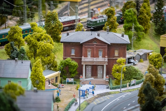 A detailed model railway scene with a prominent brick building surrounded by lush green trees. A curved road in the foreground features tiny figures and a signpost. Trains are visible in the background, traveling on tracks elevated by poles among more greenery.