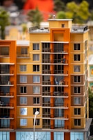 A detailed model of a modern apartment building consisting of multiple stories. The building features colorful exterior walls in shades of orange and beige, numerous balconies, and windows with some showing curtains. There's a figure on a balcony engaged in an activity like sitting or standing.