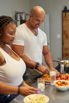 A man and a woman are preparing food in a kitchen. The man is slicing vegetables on a cutting board while the woman stands nearby holding a glass with a straw. A bowl of colorful tomatoes and a plate of sliced potatoes are on the counter. Both appear engaged and smiling, enjoying the activity.