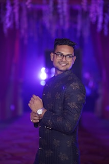 Indian male model in purple-rimmed eyeglasses smiling confidently against a soft pink background