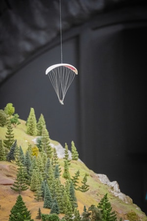 A diorama displays a miniature paraglider suspended in mid-air above a small, detailed landscape of a forested hillside. The trees are carefully crafted to resemble a variety of evergreen species, set against a backdrop of rocky terrain.