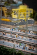 A detailed miniature model scene of a train station with several tiny figurines of people on the platforms. The scene includes visible train tracks, platforms, electrical poles, street lights, benches, and signs. In the background, there are model buildings including a yellow industrial or office building and construction equipment such as cranes.