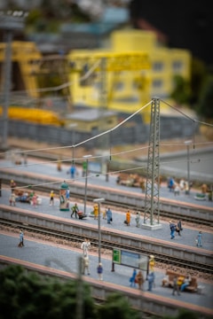 A detailed miniature model scene of a train station with several tiny figurines of people on the platforms. The scene includes visible train tracks, platforms, electrical poles, street lights, benches, and signs. In the background, there are model buildings including a yellow industrial or office building and construction equipment such as cranes.