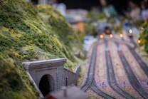 A detailed model train set with tracks curving through a realistic landscape. The foreground features a stone tunnel embedded in a lush, green hillside. The background shows multiple railway lines with small lights, simulating a busy railway scene. The focus is on the intricate details of the model landscape and railway tracks.