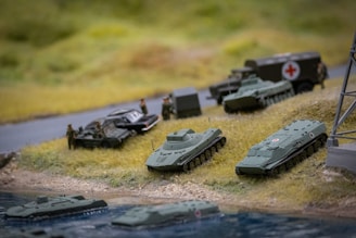 Several miniature military vehicles, including tanks and an armored personnel carrier with a red cross on it, are positioned on a grassy terrain near a body of water. There are a few miniature figures representing soldiers alongside the vehicles. The setting resembles a diorama depicting a military scene.