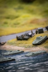 A miniature diorama depicting military vehicles near a body of water. Several model amphibious tanks are in the water, approaching a grassy shore where other military vehicles and small figures are set up, likely representing soldiers.