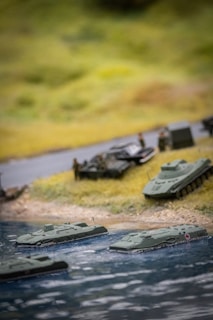 A miniature diorama depicting military vehicles near a body of water. Several model amphibious tanks are in the water, approaching a grassy shore where other military vehicles and small figures are set up, likely representing soldiers.