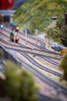 A detailed model train set with multiple tracks, featuring small trains in motion. The scene includes lush green hills in the background, tracks curving and intertwining, and miniature signal lights scattered throughout. The setting appears meticulously crafted, displaying lifelike terrain and vegetation.