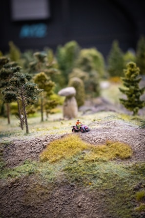 A miniature landscape featuring a small terrain with varying textures, including grass patches and dirt. A toy figure riding an all-terrain vehicle can be seen navigating through the hilly area. Surrounding the vehicle are tiny, detailed model trees enhancing the sense of a natural environment.
