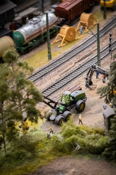 The image depicts a detailed miniature model scene featuring a railway setting with tracks, a green tractor-like vehicle carrying logs, and a few miniature figures. In the background, there are trains and some yellow cable spools, hinting at a construction or industrial environment. The foreground is filled with greenery, suggesting a rural or outdoor setting.