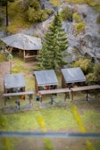 A miniature scene depicting an outdoor setting with several small wooden structures resembling market stalls or shelters. There is a larger wooden gazebo with a pitched roof in the background, surrounded by trees and vegetation. The scene includes miniature figures of people under the stalls and gazebo. A tall pine tree and a vintage-style street lamp are also part of the scene, with rocky cliffs and greenery in the background.