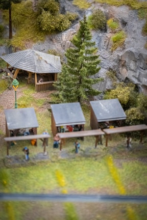 A miniature scene depicting an outdoor setting with several small wooden structures resembling market stalls or shelters. There is a larger wooden gazebo with a pitched roof in the background, surrounded by trees and vegetation. The scene includes miniature figures of people under the stalls and gazebo. A tall pine tree and a vintage-style street lamp are also part of the scene, with rocky cliffs and greenery in the background.