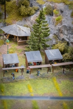 A miniature scene depicting an outdoor setting with several small wooden structures resembling market stalls or shelters. There is a larger wooden gazebo with a pitched roof in the background, surrounded by trees and vegetation. The scene includes miniature figures of people under the stalls and gazebo. A tall pine tree and a vintage-style street lamp are also part of the scene, with rocky cliffs and greenery in the background.