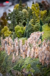 A miniature landscape features detailed rocky formations and lush green trees. Tall, slender rock pillars rise from a grassy terrain, surrounded by various types of dense, fluffy trees resembling a forest. The scene captures a realistic diorama of a natural environment with intricate attention to texture and color.