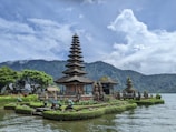 A majestic traditional temple is situated on a small island surrounded by water, with lush green gardens and ornate statues. In the background, there are rolling hills and a partly cloudy sky, creating a serene and picturesque setting.