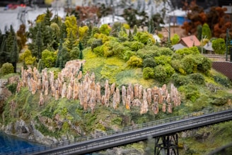 Miniature diorama featuring a lush, green landscape with dense foliage and tall, stone-like structures on a hill. The scene includes a small section of water and a bridge in the foreground, with tiny buildings visible in the background.