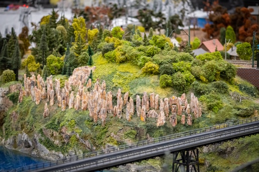 Miniature diorama featuring a lush, green landscape with dense foliage and tall, stone-like structures on a hill. The scene includes a small section of water and a bridge in the foreground, with tiny buildings visible in the background.