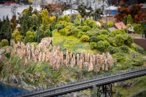 Miniature diorama featuring a lush, green landscape with dense foliage and tall, stone-like structures on a hill. The scene includes a small section of water and a bridge in the foreground, with tiny buildings visible in the background.