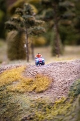 A miniature scene depicts a figure riding a small vehicle across a textured landscape with patches of green and yellow grass. Tall, fuzzy trees are in the background, contributing to a scaled-down, crafted appearance.