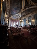 A lavish and opulent room with ornate ceiling paintings and intricate gold detailing throughout. The room features luxurious red and gold upholstered chairs and sofas, with elegant chandeliers hanging from the ceiling. Large draped windows allow natural light to illuminate the rich interior, creating a warm and sophisticated atmosphere.