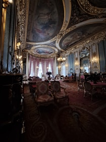 A lavish and opulent room with ornate ceiling paintings and intricate gold detailing throughout. The room features luxurious red and gold upholstered chairs and sofas, with elegant chandeliers hanging from the ceiling. Large draped windows allow natural light to illuminate the rich interior, creating a warm and sophisticated atmosphere.